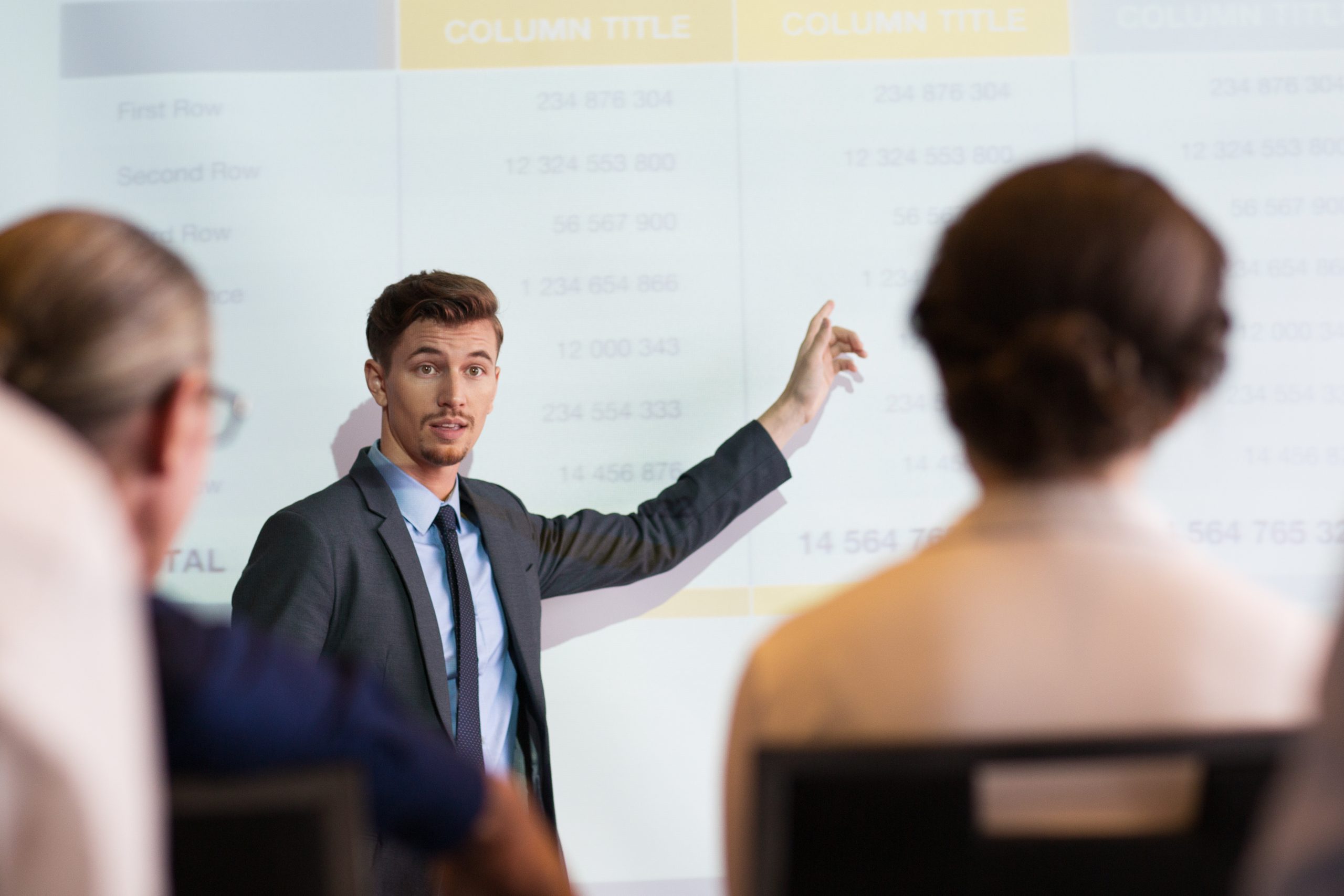 Serious middle-aged businessman standing at projection screen with table and pointing to it while looking and explaining ideas to audience seen partly